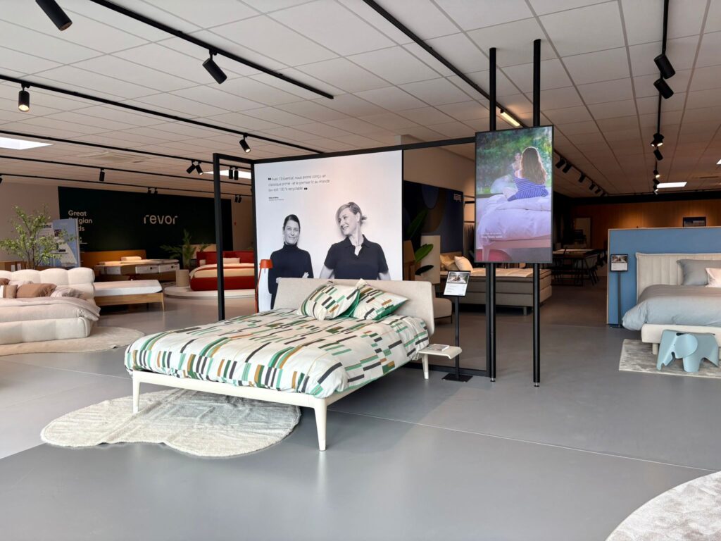 Showroom Revor avec lits exposés, écrans affichant deux femmes, mobilier design et accessoires de décoration intérieure