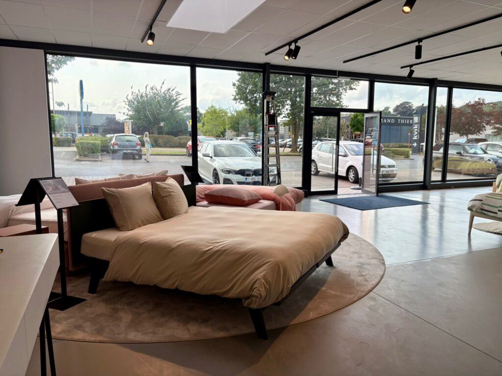Lit design avec draps beige exposé dans un showroom de mobilier avec baies vitrées donnant sur le parking