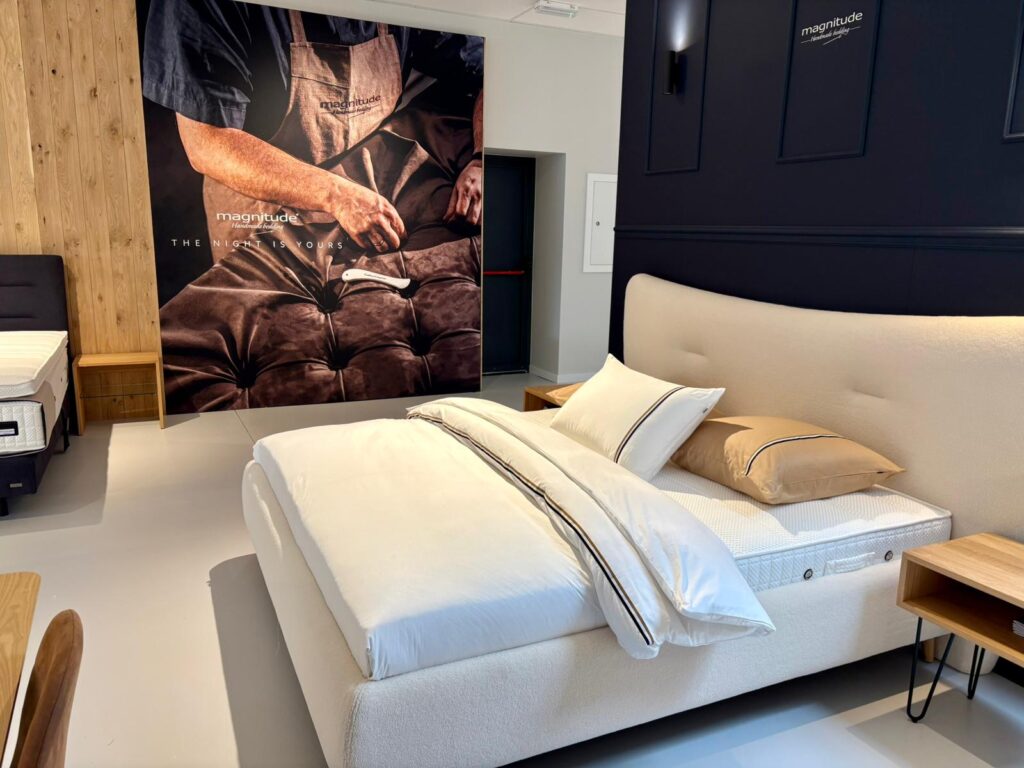 Lit Magnitude avec matelas blanc, draps beige et affiche publicitaire The night is yours en showroom