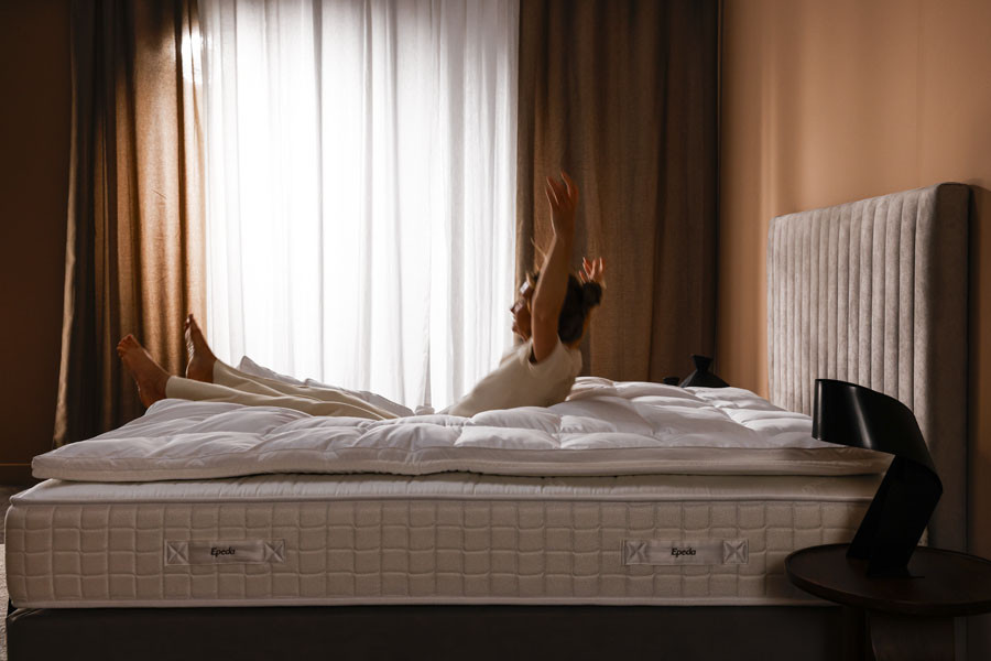 Femme s'étirant sur un surmatelas Epeda Sublime dans une chambre lumineuse avec rideaux beige